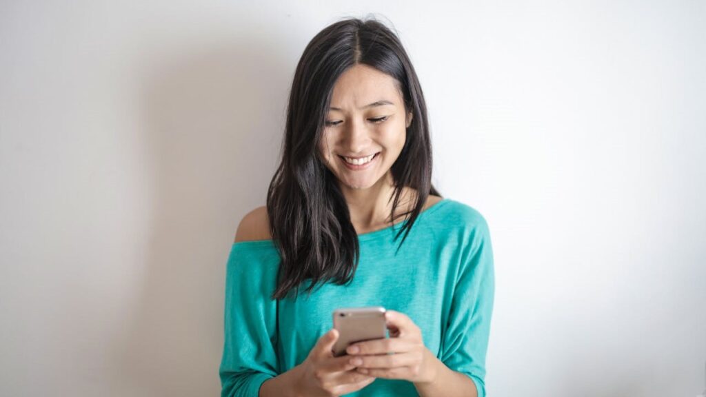 Vrouw glimlacht terwijl ze een anonieme sms leest op haar smartphone in een turquoise trui