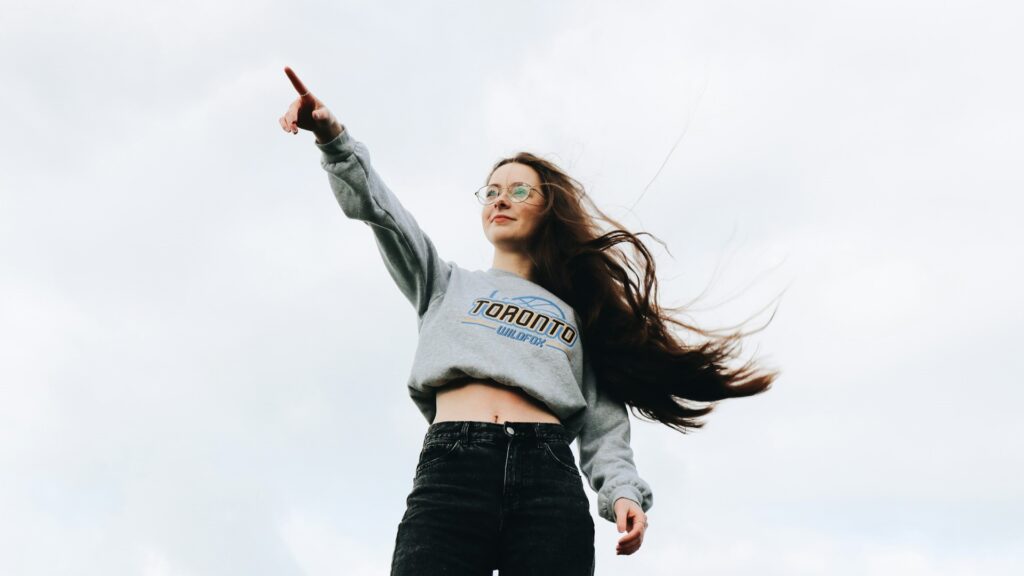 Zelfverzekerde jonge vrouw met lang haar wijst omhoog in de lucht, een symbool van groeiend zelfvertrouwen.